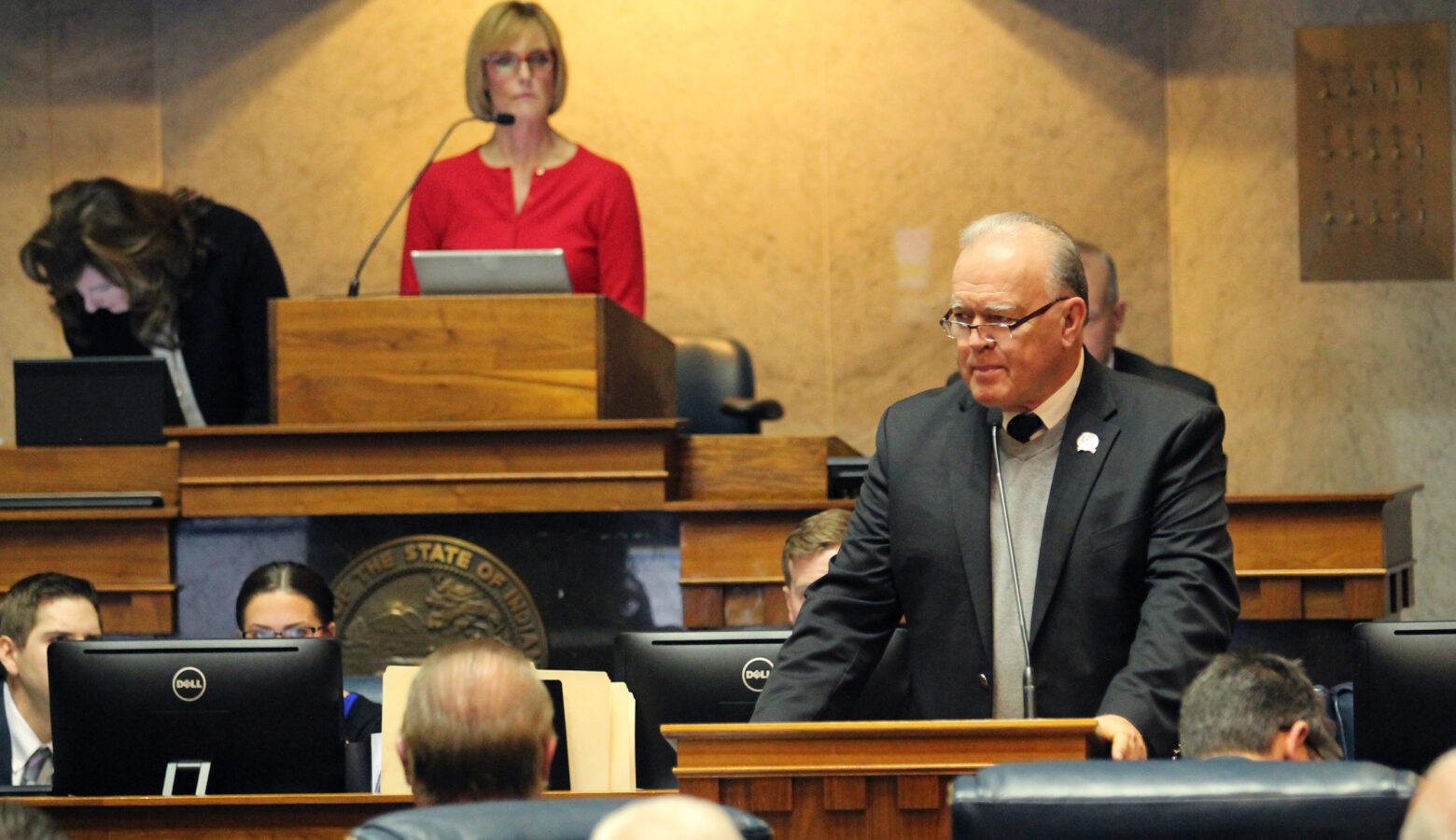 Senator Jim Buck stands at a podium on the Senate floor. He is a White man, balding with white and grey hair. He wears a suit and glasses.